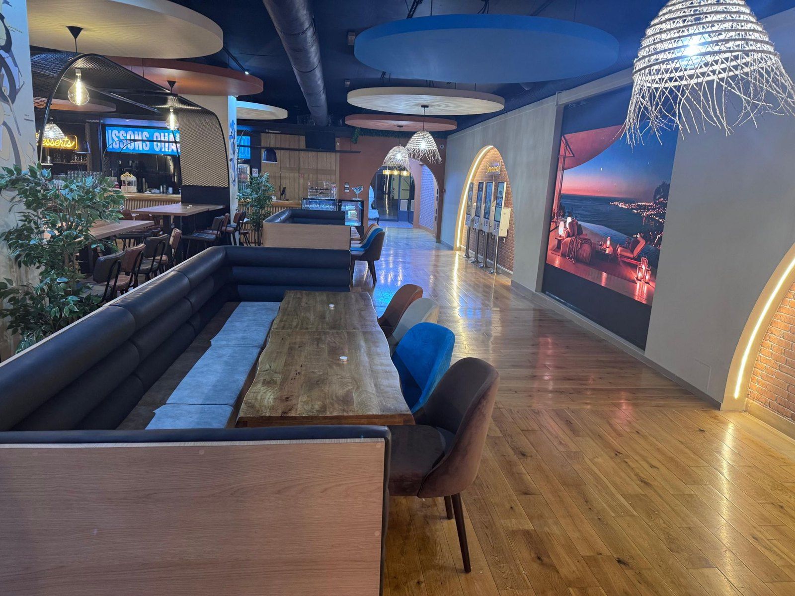 Une longue table en bois brut, sa banquette et ses chaises de velours sous les suspensions tressées ; au fond, le comptoir des desserts et les bornes de commande